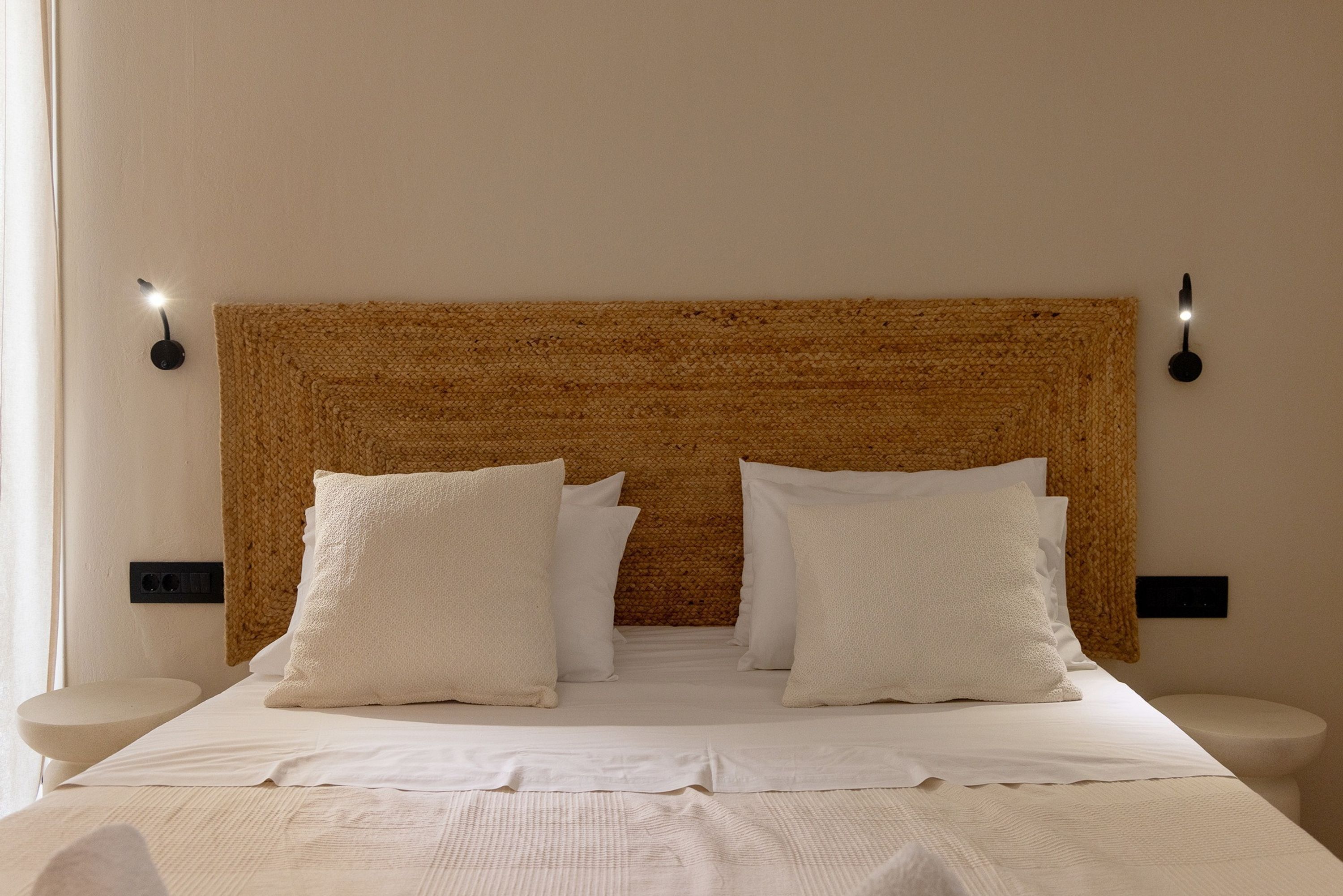 Een net opgemaakt bed met witte lakens en kussens, een gestructureerd geweven hoofdbord en nachtkastjes met zwart wandlampjes.