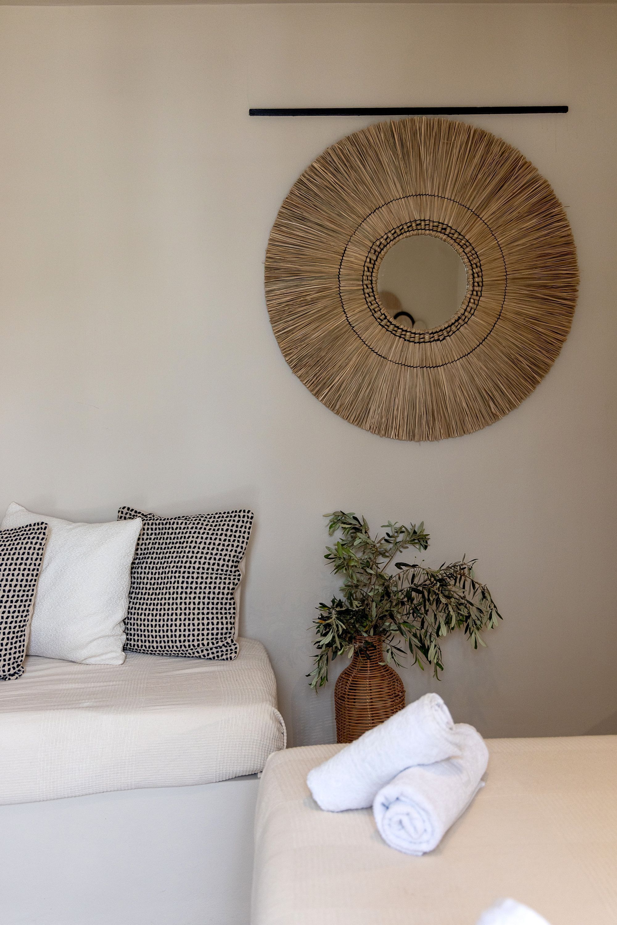 A room with a bed, two pillows, a woven wall decoration, a potted plant, and a rolled towel on a bedside table.