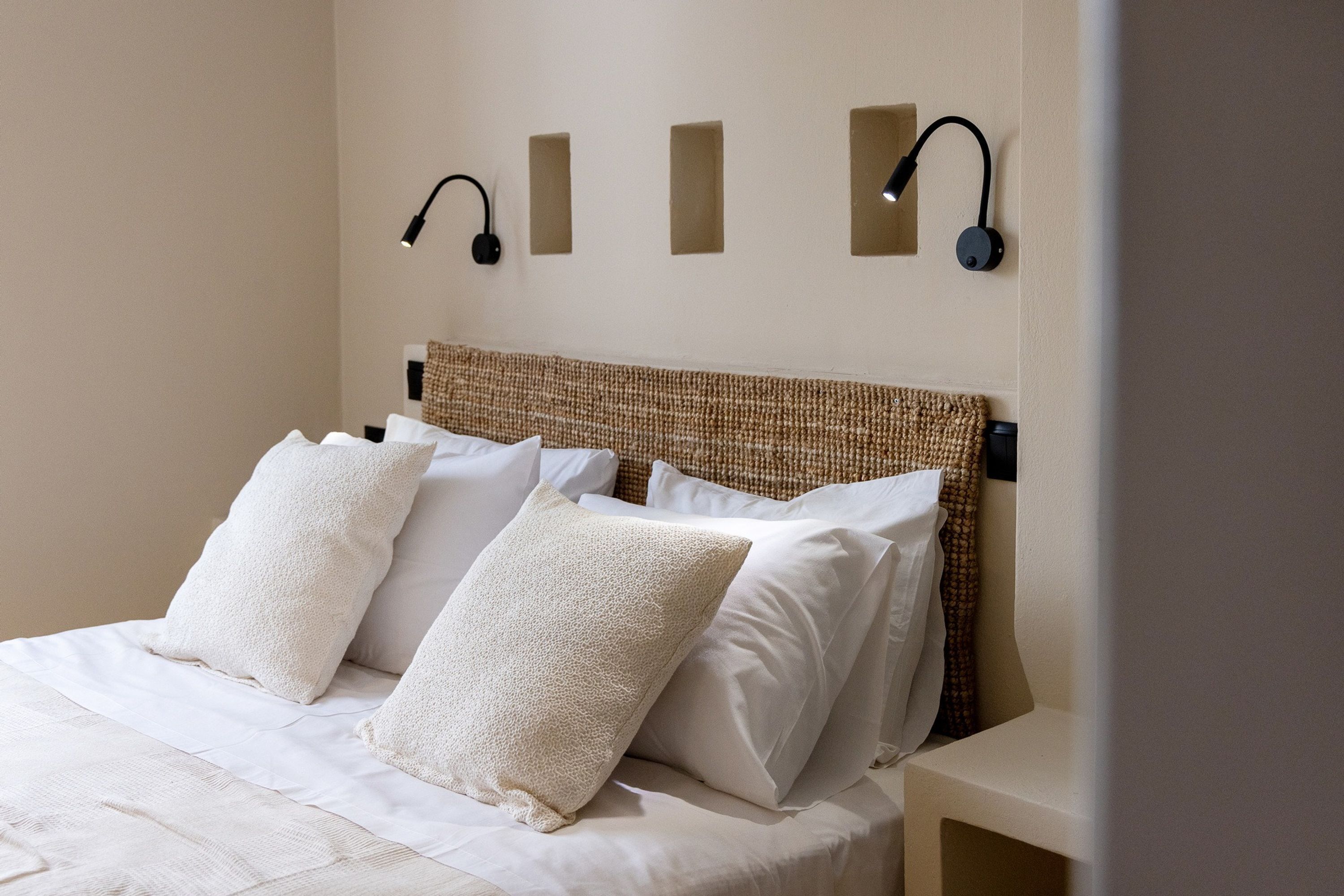 Una cama bien hecha con sábanas y almohadas blancas, un cabecero tejido y dos lámparas fijadas a la pared.