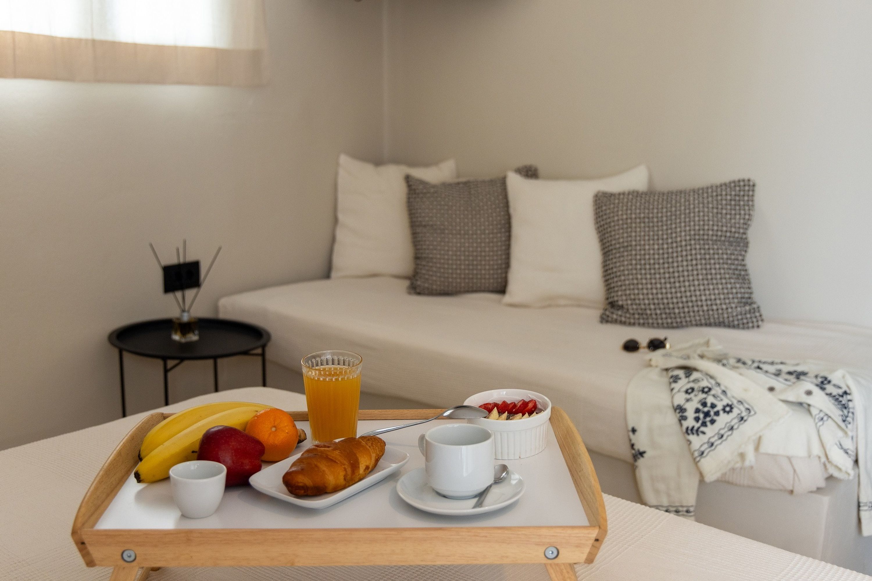 A breakfast tray with fruits, a croissant, and a glass of orange juice on a wooden table in a hotel room with a bed and pillows in the background.