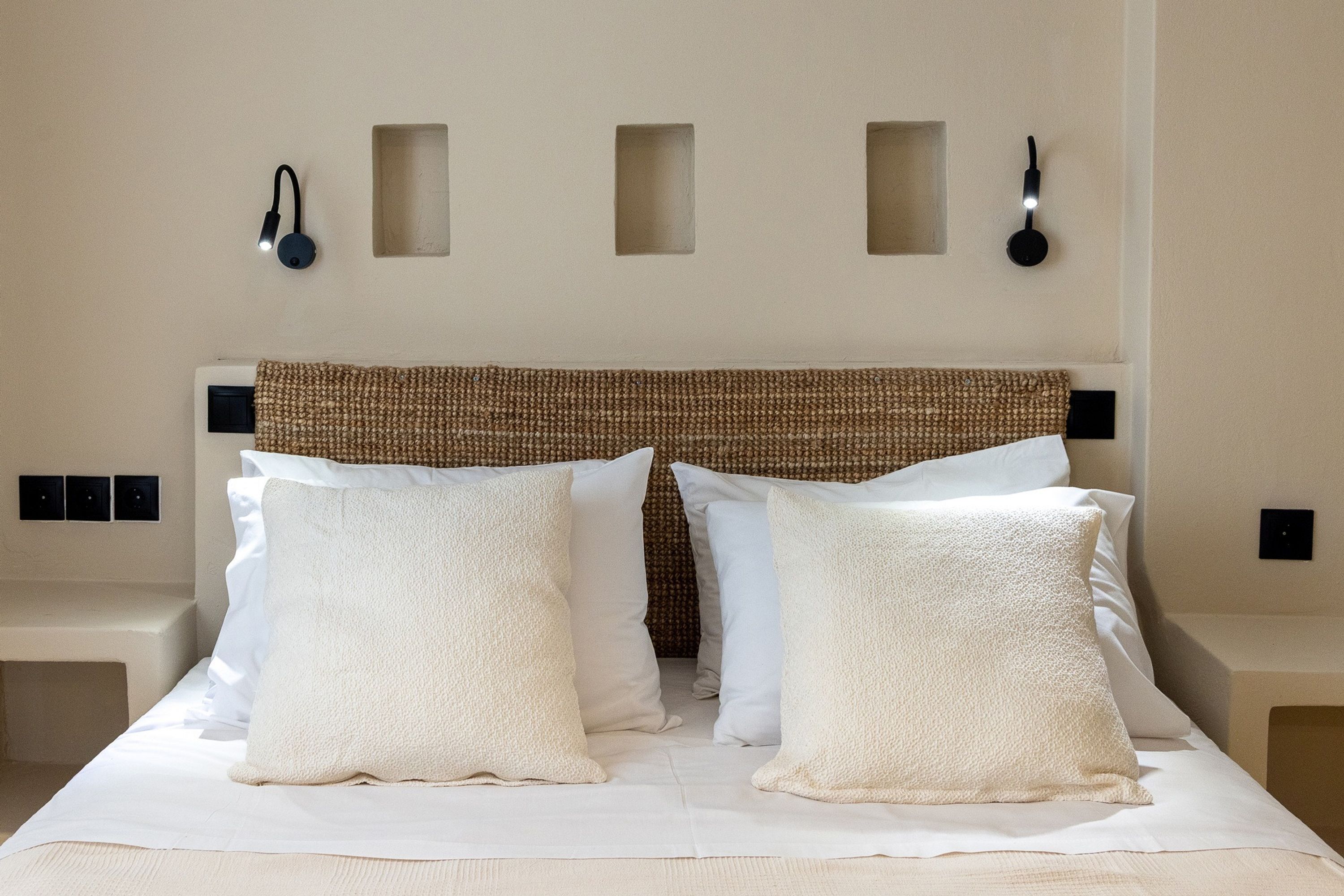 A neatly made bed with white linens and two large pillows, a woven headboard, and two wall-mounted light fixtures.