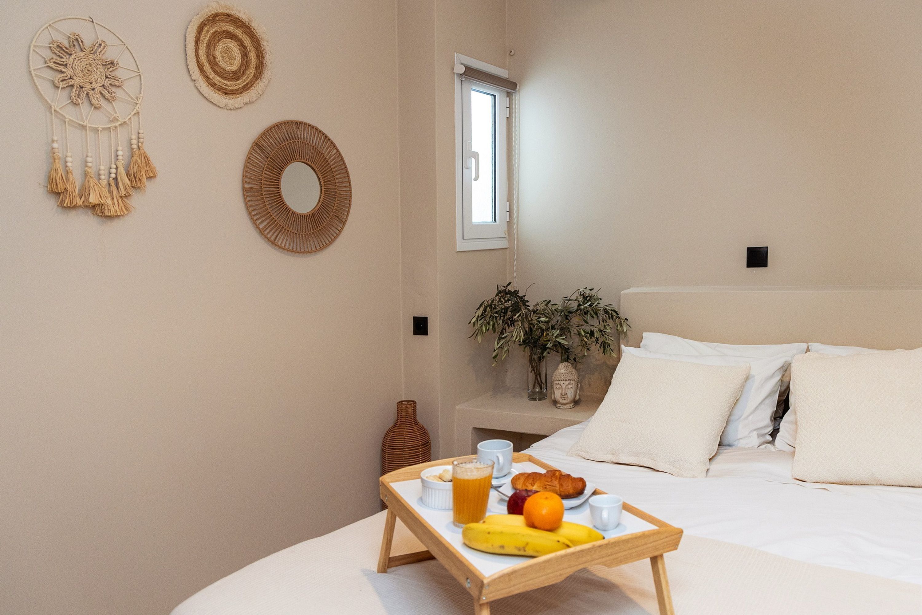 Un lit bien fait, avec du linge de lit blanc, une table de chevet ornée d’un vase et d’une plante, et un plateau de petit-déjeuner avec des fruits et une boisson posé sur le lit.
