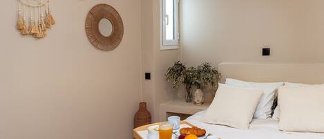 Un lit bien fait, avec du linge de lit blanc, une table de chevet ornée d’un vase et d’une plante, et un plateau de petit-déjeuner avec des fruits et une boisson posé sur le lit.