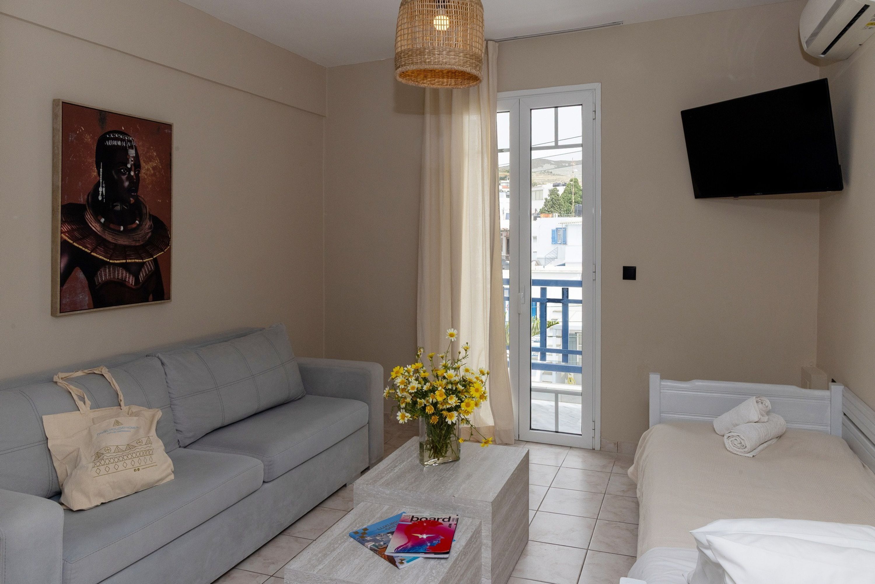 Habitación de hotel con sofá gris, cama blanca, televisor de pantalla plana, un cuadro en la pared y ventana con vistas a edificios.