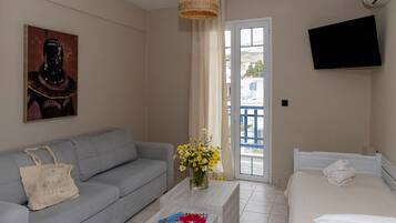 Habitación de hotel con sofá gris, cama blanca, televisor de pantalla plana, un cuadro en la pared y ventana con vistas a edificios.