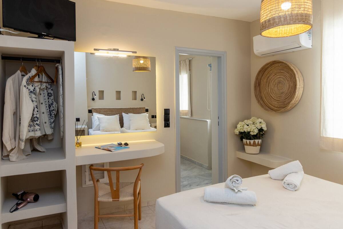 A modern hotel room with a bed, a desk with a chair, a wardrobe, a mirror, and a shower area.
