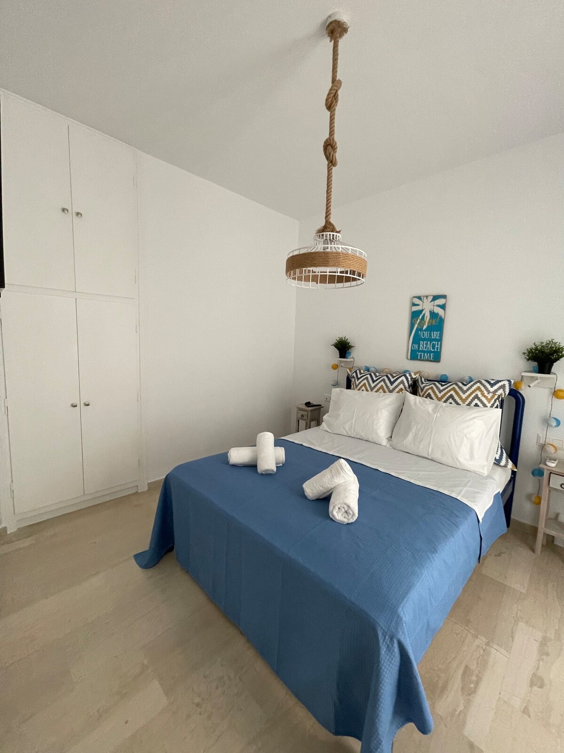 A neatly made bed with a blue bedspread and white pillows, a hanging rattan light fixture, and a wall poster.