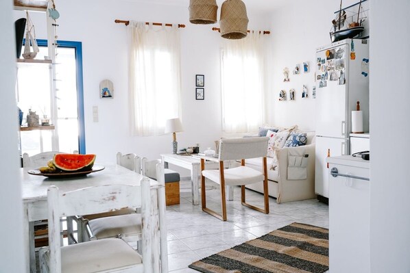 A bright, well-lit kitchen with white cabinetry, a dining table with chairs, and a refrigerator with magnets and pictures. - Menmar House in Tinos (Tinos)