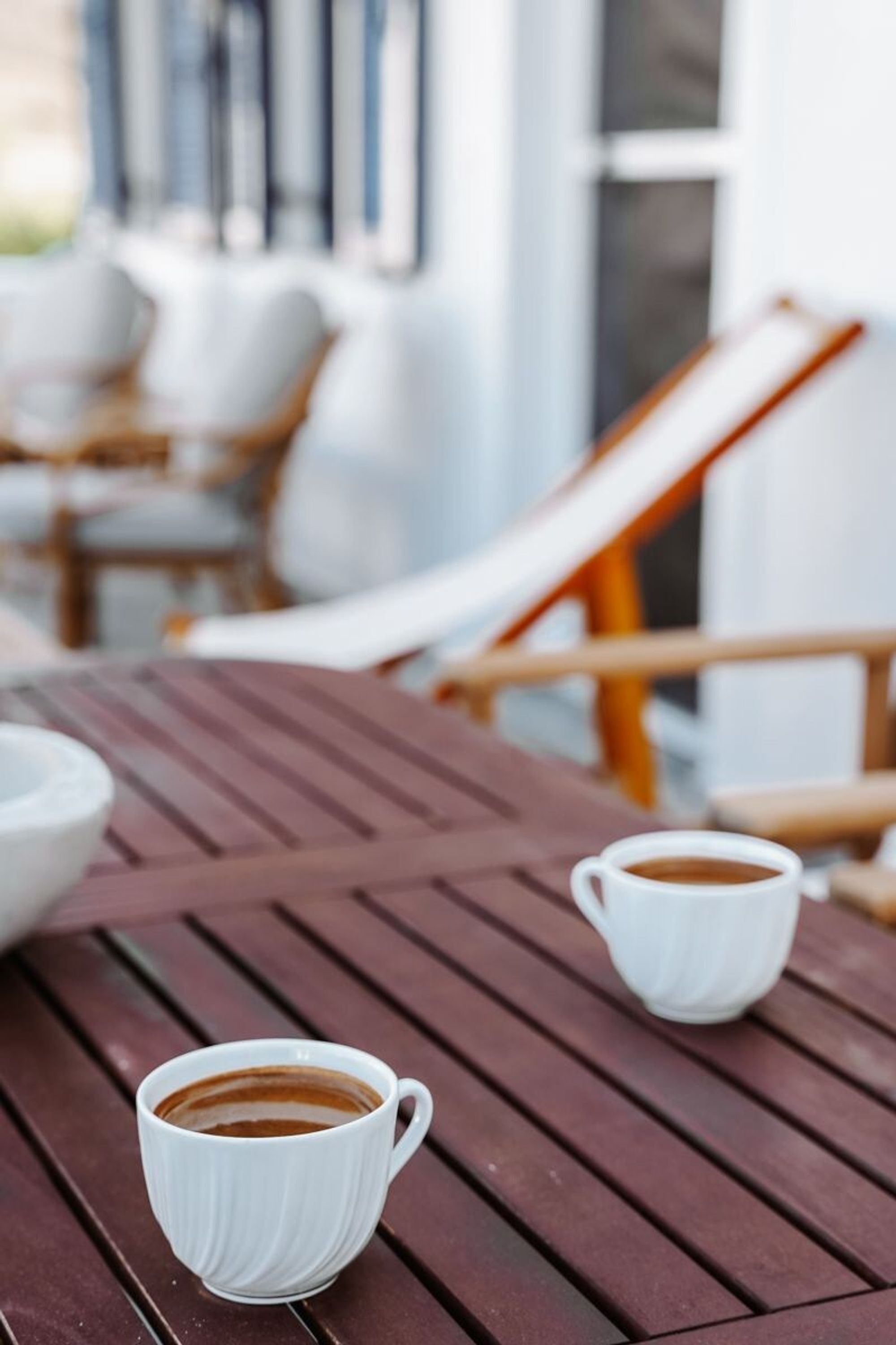 A wooden table with two white cups of coffee, a white bowl, and a wooden chair with a cushion.