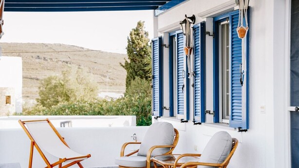 Une terrasse avec des chaises en osier et un fauteuil en toile, des murs blancs, des volets bleus et une vue sur les collines.