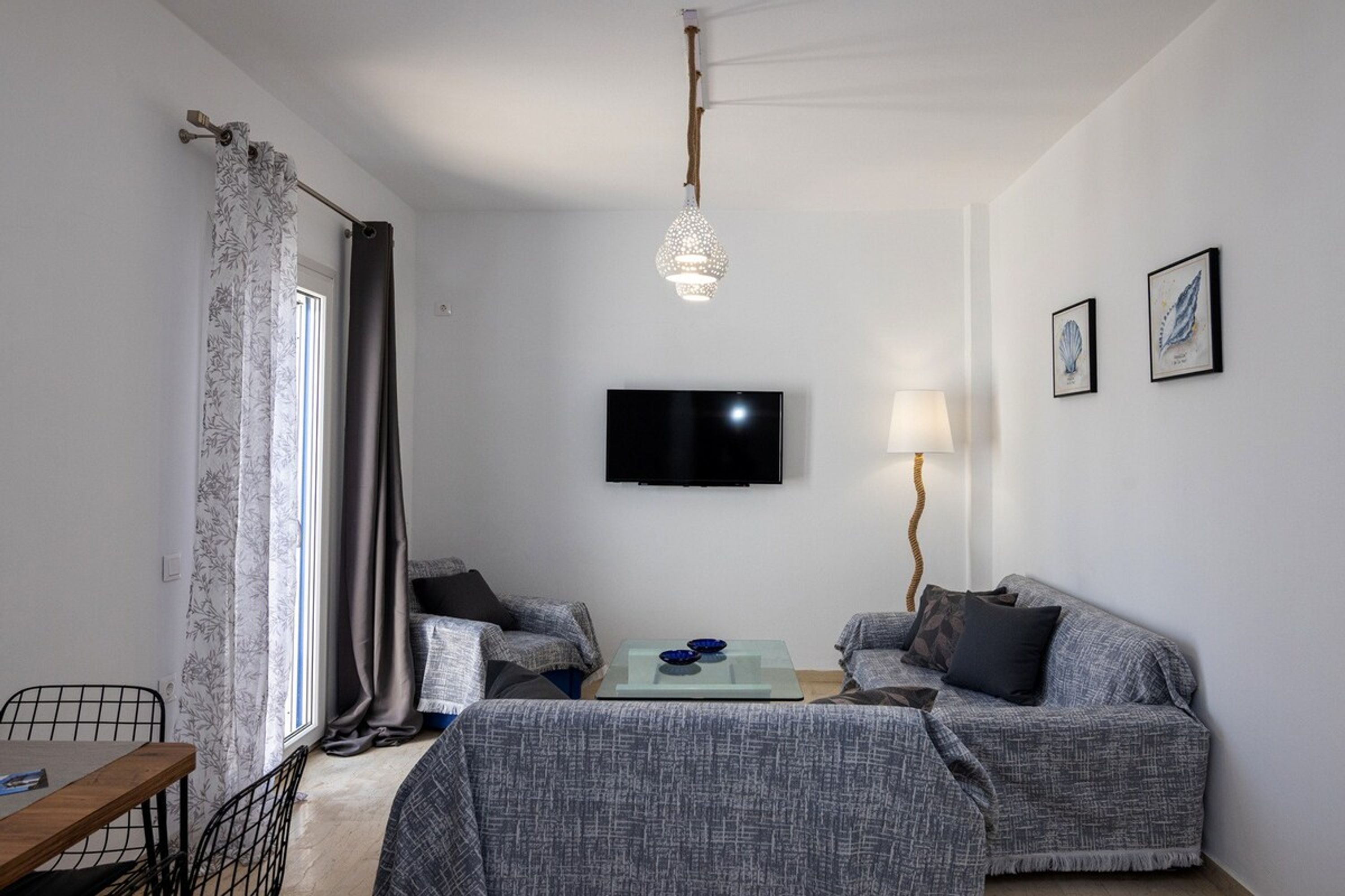 A living room with two grey sofas, a glass coffee table, a flat-screen TV, a hanging light fixture, and a window with curtains.