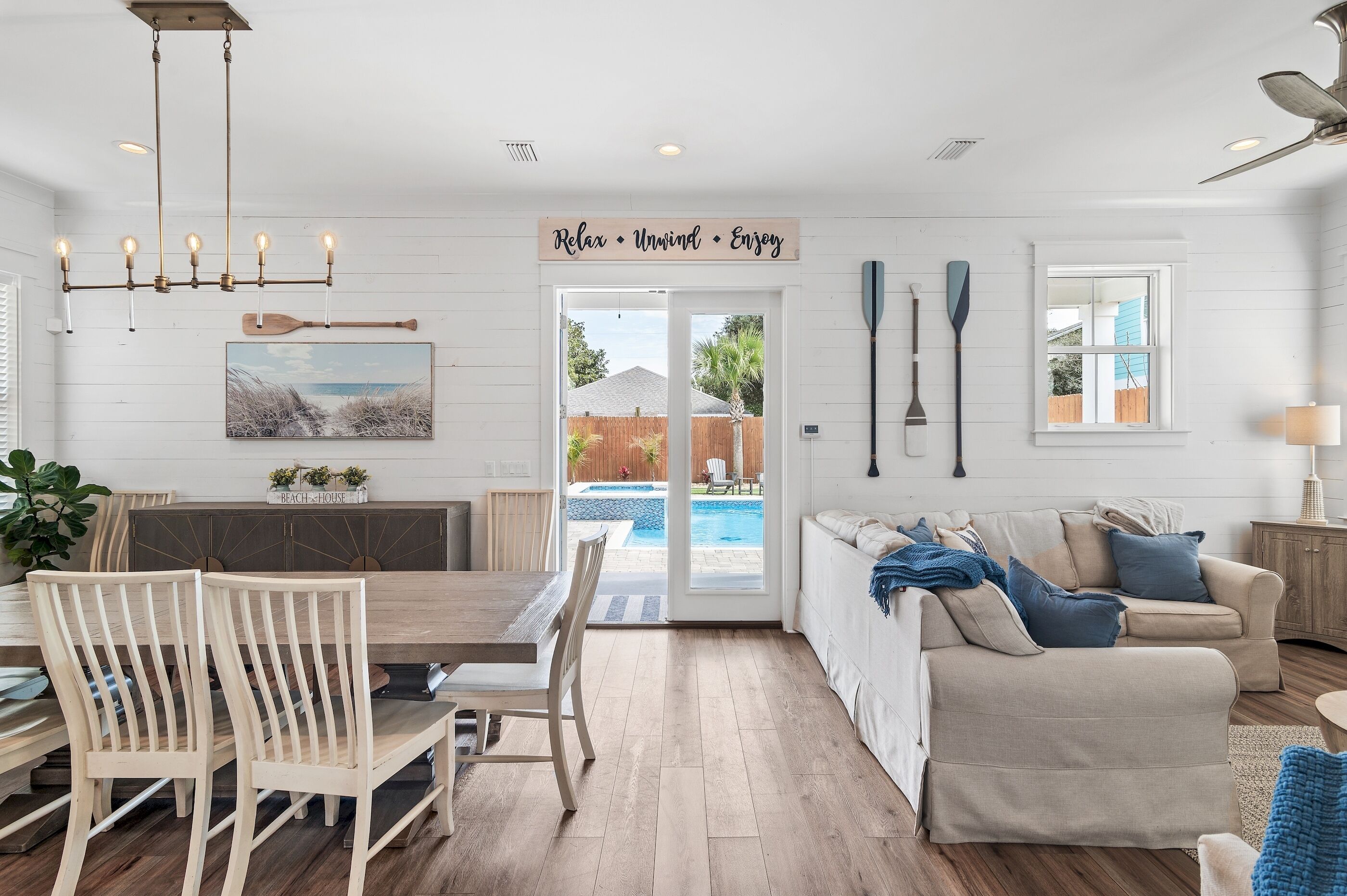 A spacious living area with a dining table, chairs, a sofa, and a view of a swimming pool through glass doors.