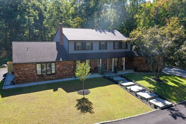 A two-story house with a brick facade, a grey roof, and a well-maintained lawn with a tree and stepping stones. - Spacious 5-bedroom house in the woods in Cold Spring just 5 minutes from town (Cold Spring)