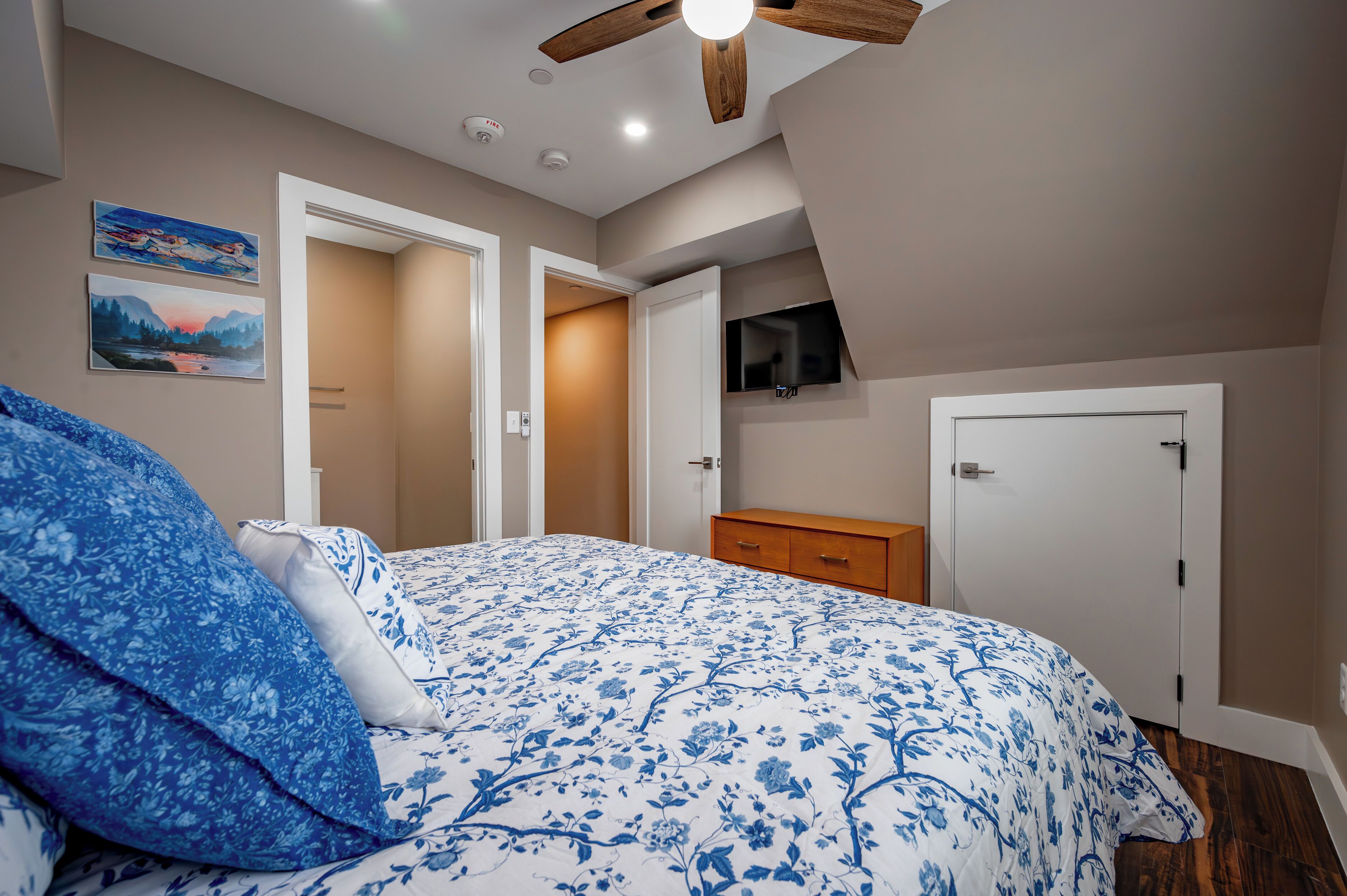 A bedroom with a bed, a wooden dresser, a TV mounted on the wall, and a ceiling fan.