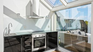 A modern kitchen with a glass enclosure, stainless steel appliances, and a view of a wicker sofa through large windows.