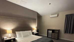 A modern hotel room with a large bed, two bedside tables with lamps, a small desk with drawers, an air conditioning unit, and a window with curtains. - Dream Hotel Bronx (Mount Vernon)