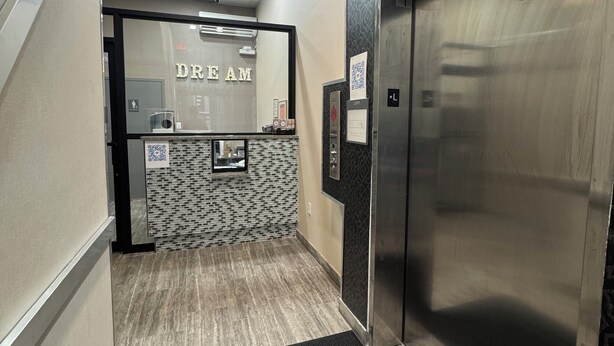 An elevator with a metallic finish, a patterned wall, and a black carpet leading to a reception area with a tiled floor and a mirror. - Dream Hotel Bronx (Mount Vernon)