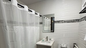 A bathroom with a shower curtain, a sink with a mirror, a toilet, and a trash bin. - Dream Hotel Bronx (Mount Vernon)