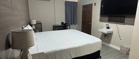 A modern hotel room with a bed, bedside lamp, desk, chair, TV, and a small sink.