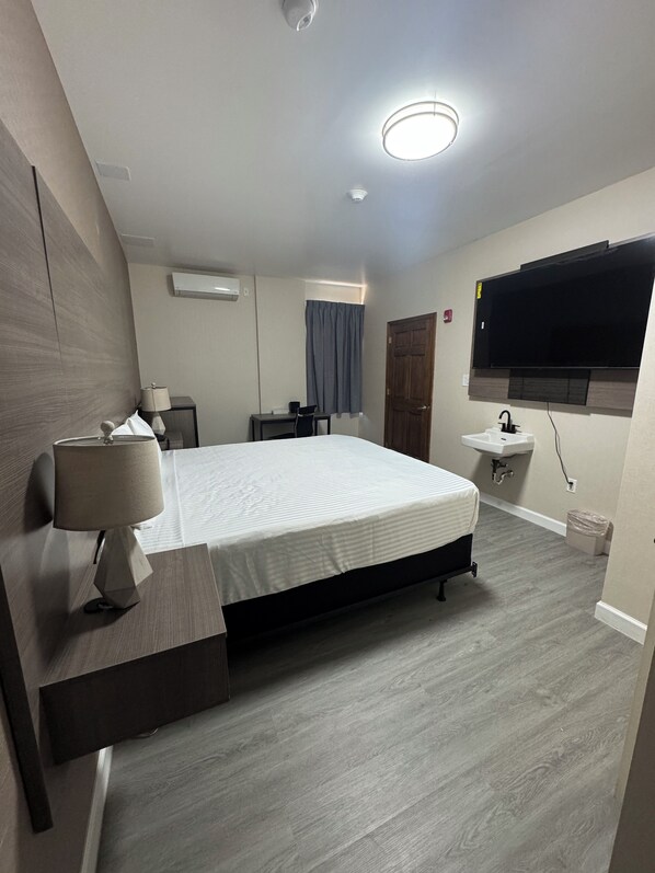 A modern hotel room with a bed, bedside lamp, desk, chair, TV, and a small sink.