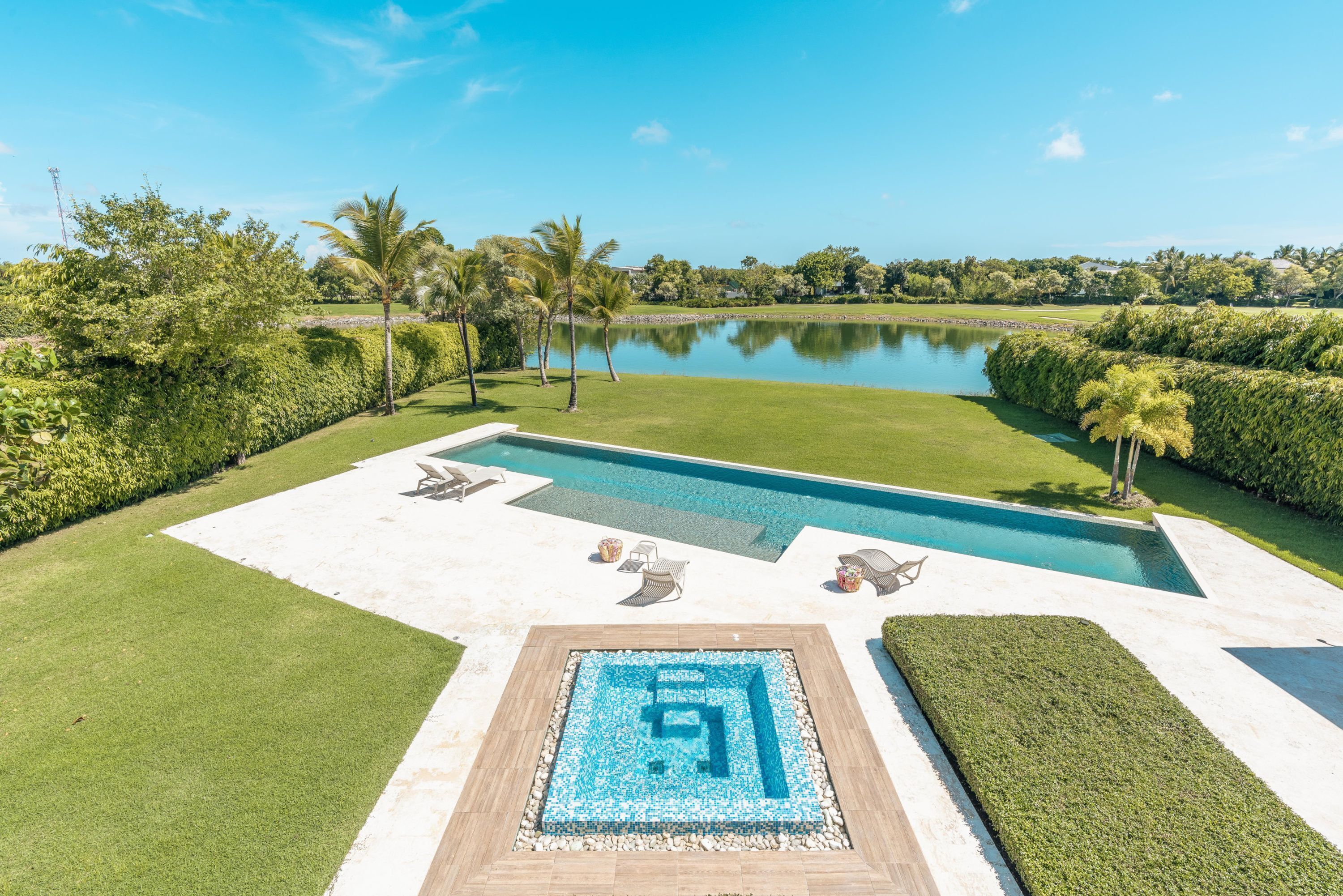A swimming pool with a geometric design, surrounded by manicured lawns and palm trees, with a view of a lake and distant buildings.