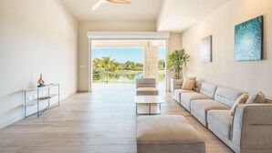 A spacious living room with a large sofa, a coffee table, and a ceiling fan. A large window offers a view of a pool and greenery.