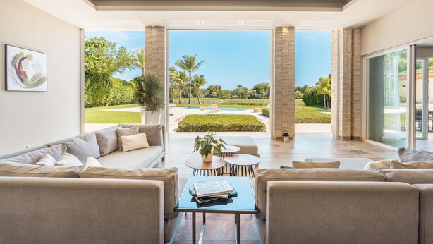 A spacious living room with a large window, a sofa, a coffee table, and a view of a well-manicured garden.
