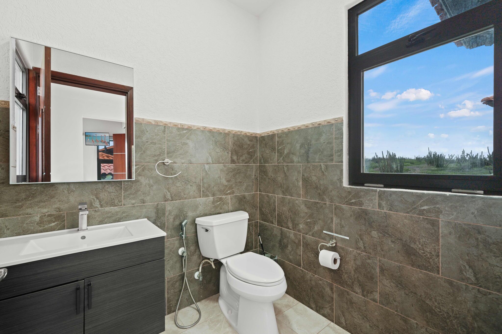 Ein Badezimmer mit einem weißen Waschbecken, einem dunklen Holzschrank, einer weißen Toilette und einem Fenster mit Blick auf Bäume.