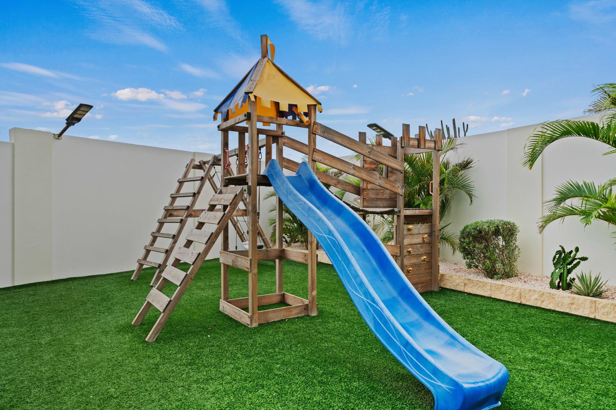 A wooden play structure with a slide and climbing area, surrounded by artificial grass and palm plants.