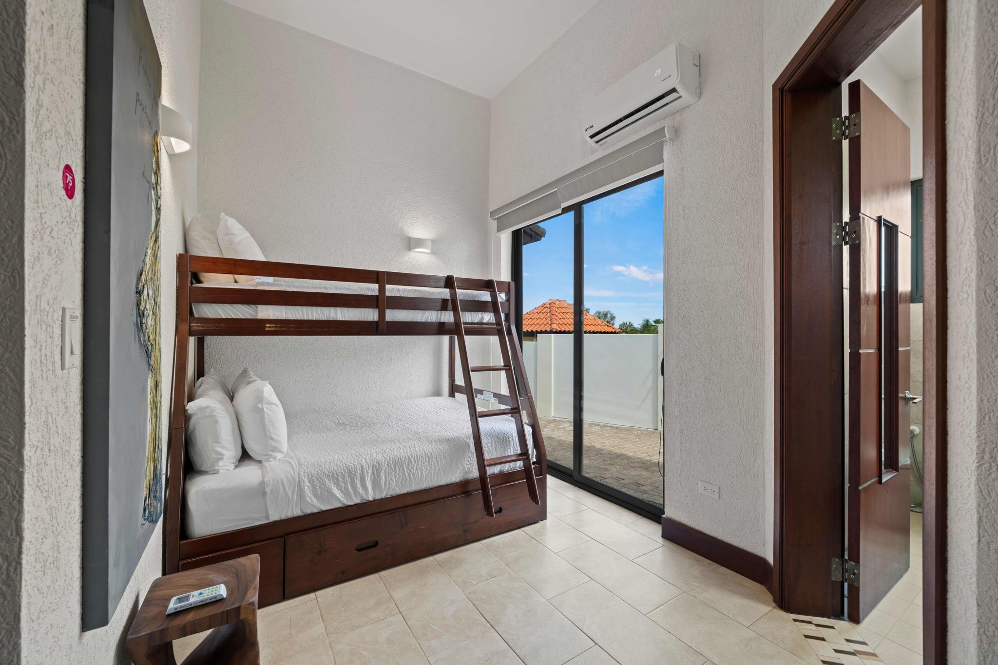 A bedroom with a bunk bed, a ladder, a small table, and a remote control. There is an air conditioning unit on the wall and a sliding glass door leading to an outdoor area.