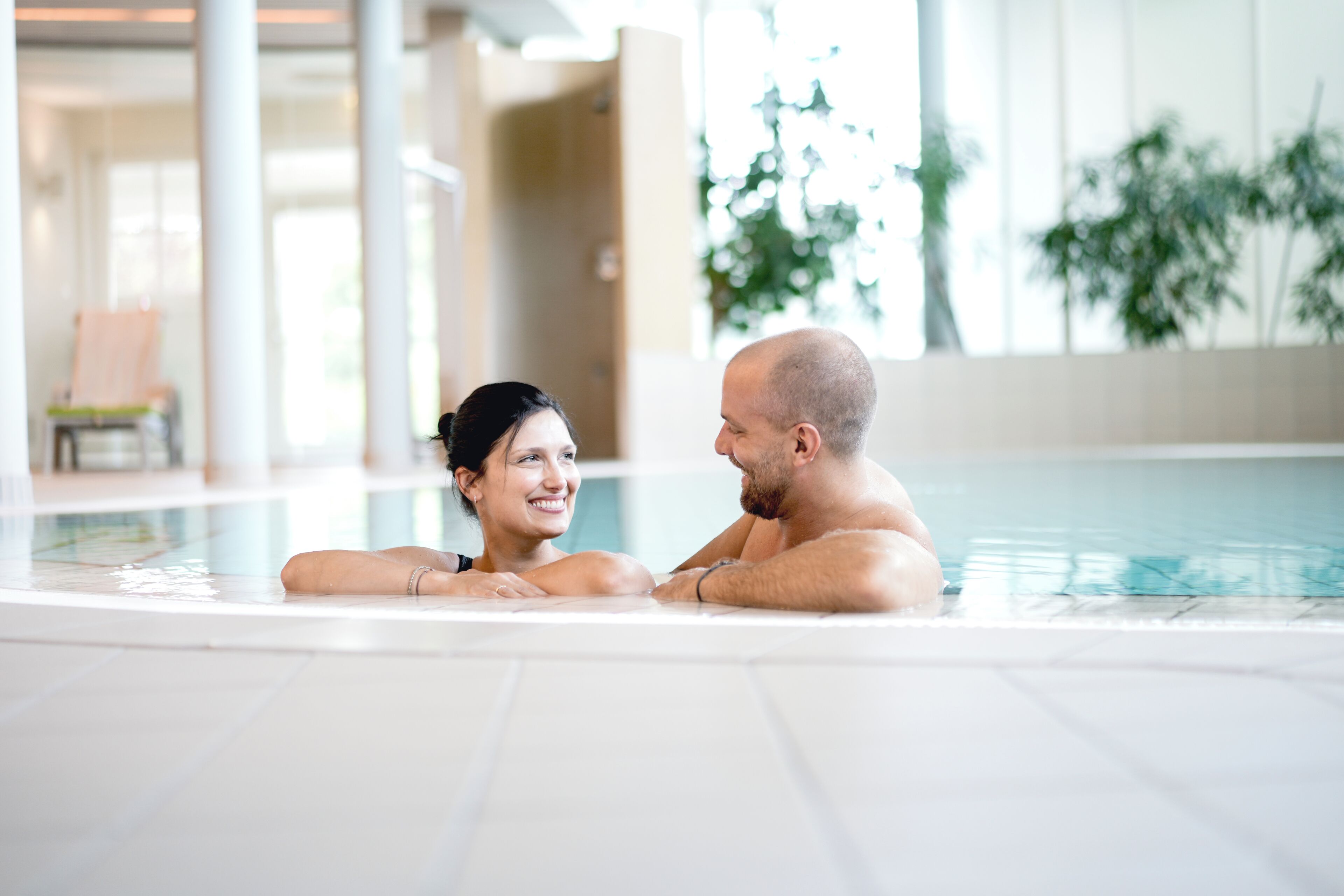Un couple dans une piscine, dans un décor intérieur moderne avec de grandes fenêtres.
