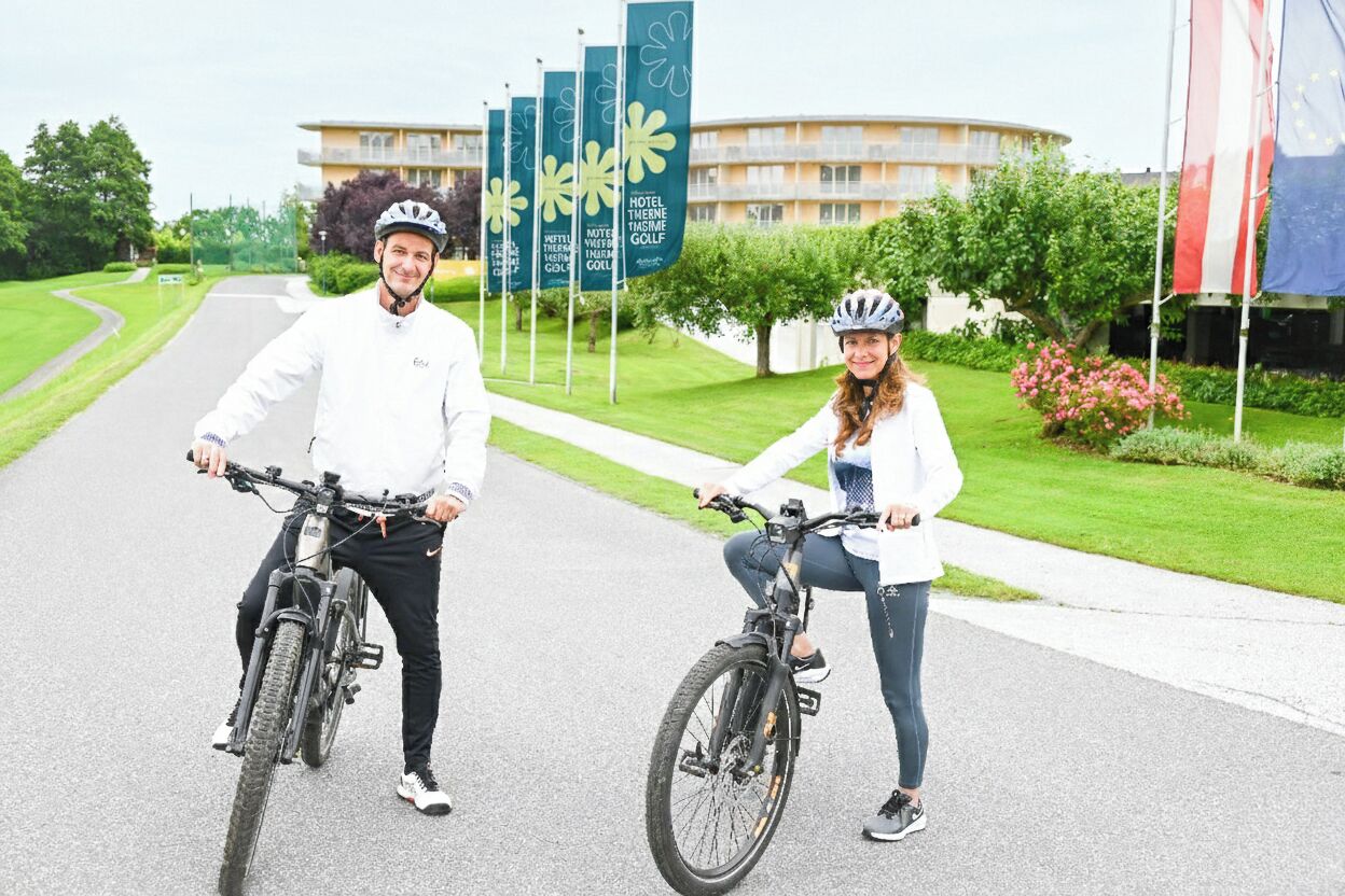 To personer på cykler foran en bygning med flag og et skilt, der står 'HOTEL THERME GOLF'.