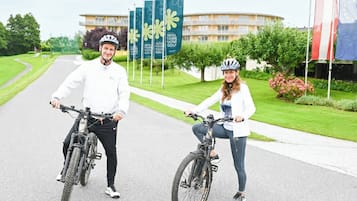 To personer på cykler foran en bygning med flag og et skilt, der står 'HOTEL THERME GOLF'.