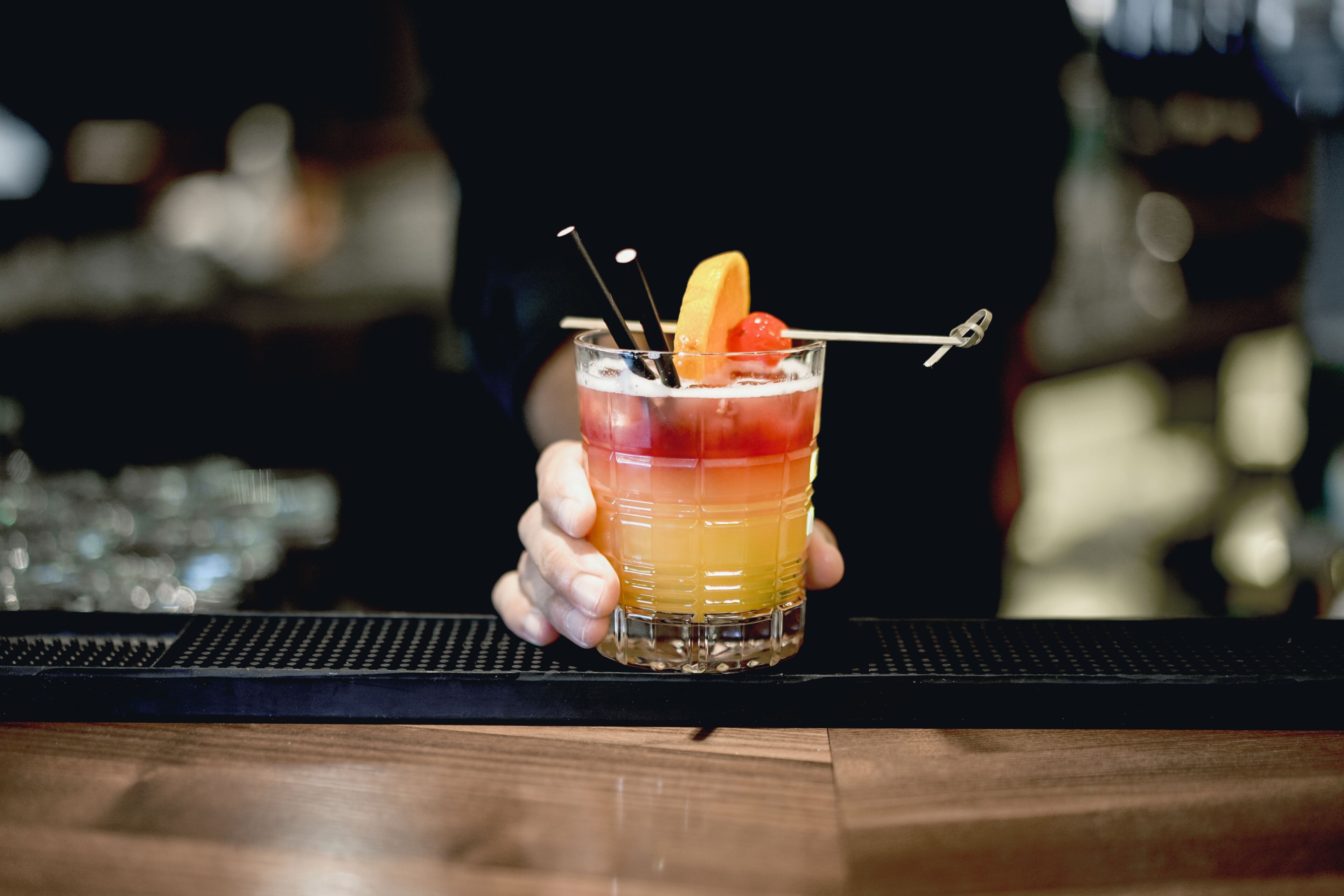 A bartender holding a layered cocktail with a cherry and lemon slice garnish.