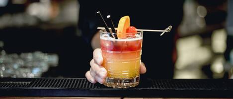 Baarimikko pitää kädessään kerroksittain valmistettua cocktailia, joka on koristeltu kirsikalla ja sitruunaviipaleella.