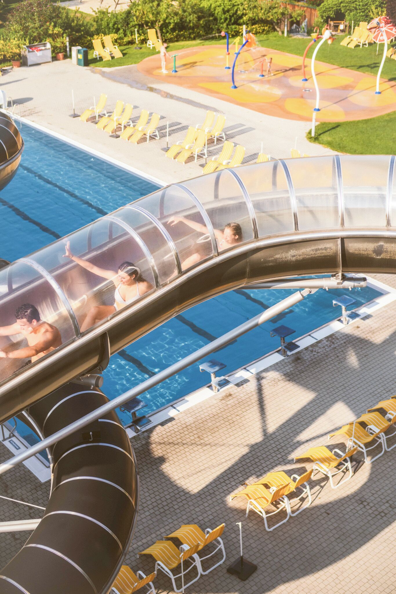 A water slide with people sliding down into a pool area, surrounded by sun loungers and a water park.