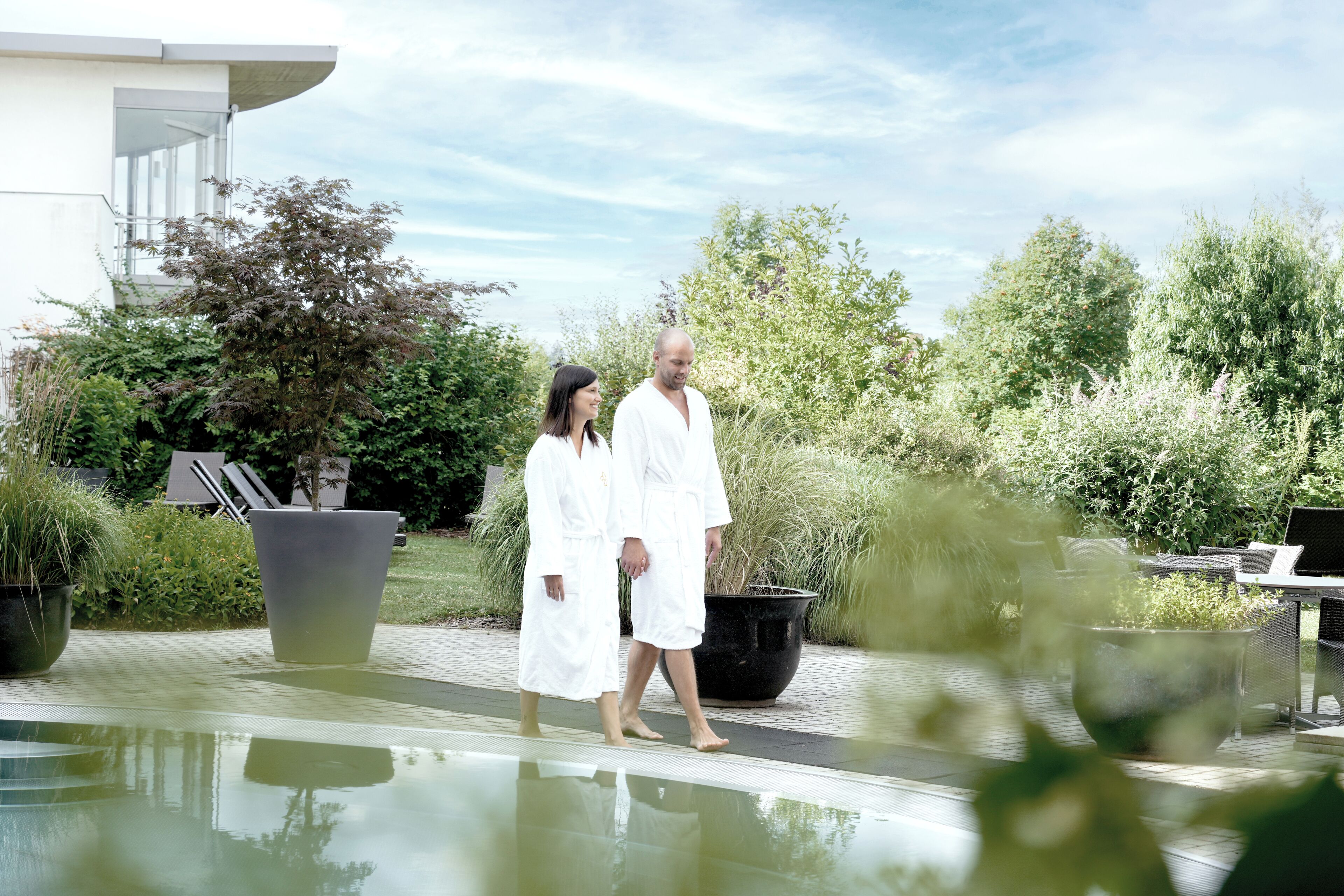 A couple walking by a pool with a modern building and lush greenery in the background.