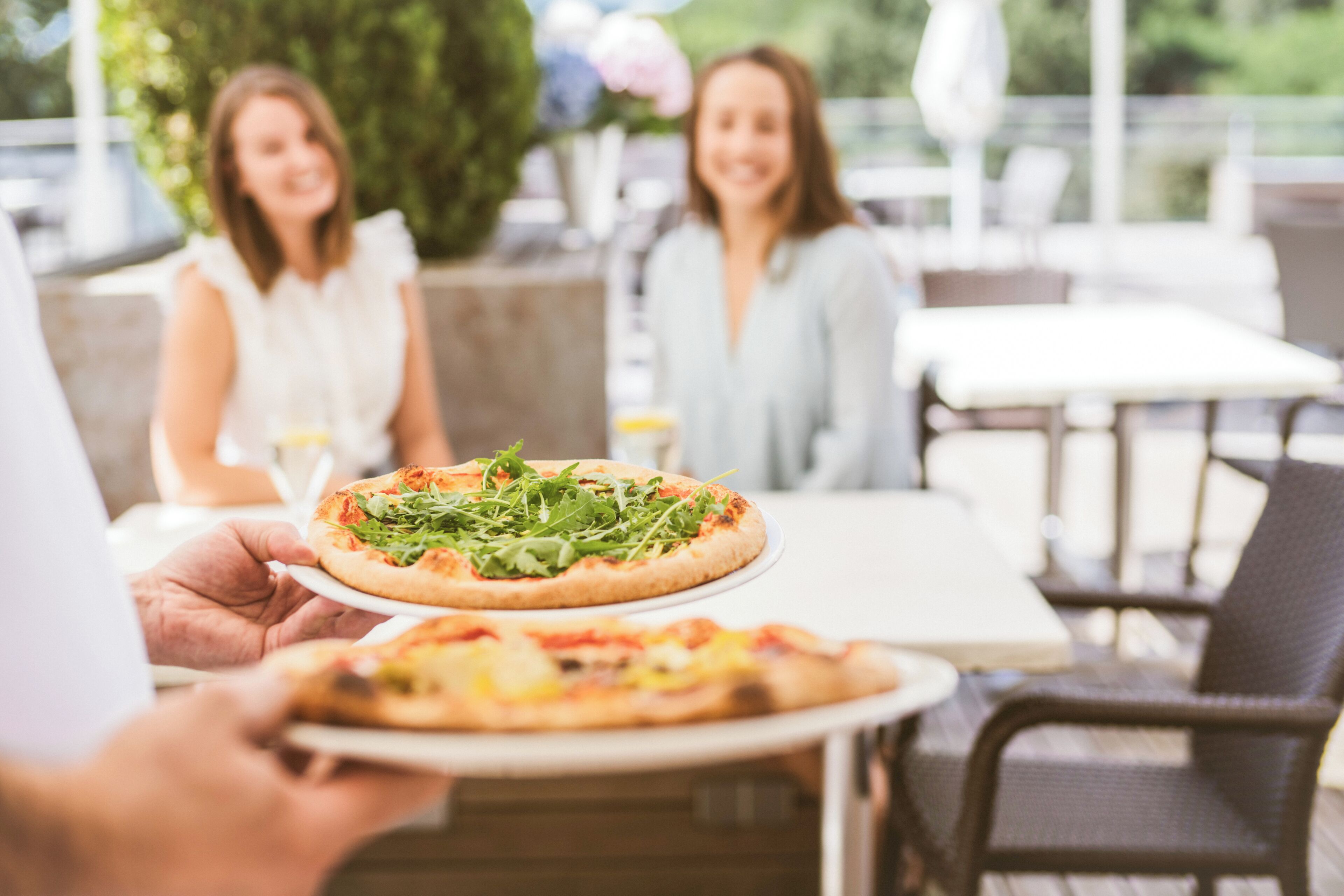 Una persona sostiene una pizza con un plato, sentada en una mesa exterior con sillas y un mantel blanco.