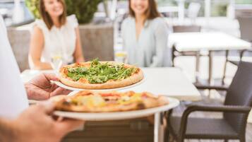 Una persona sostiene una pizza con un plato, sentada en una mesa exterior con sillas y un mantel blanco.