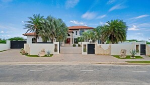 A modern two-story house with a red-tiled roof, a gated driveway, and palm trees. - Lux Villa Oasis: 5BR w Pool* Playground near Beach (Noord)