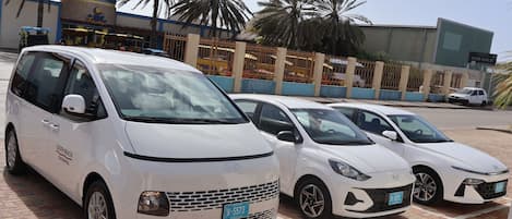 Un estacionamiento con varios vehículos blancos, palmeras y un edificio con un cartel que dice 'For Sale'.