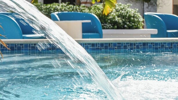 Una piscina con fuente, sillas azules y follaje al fondo.