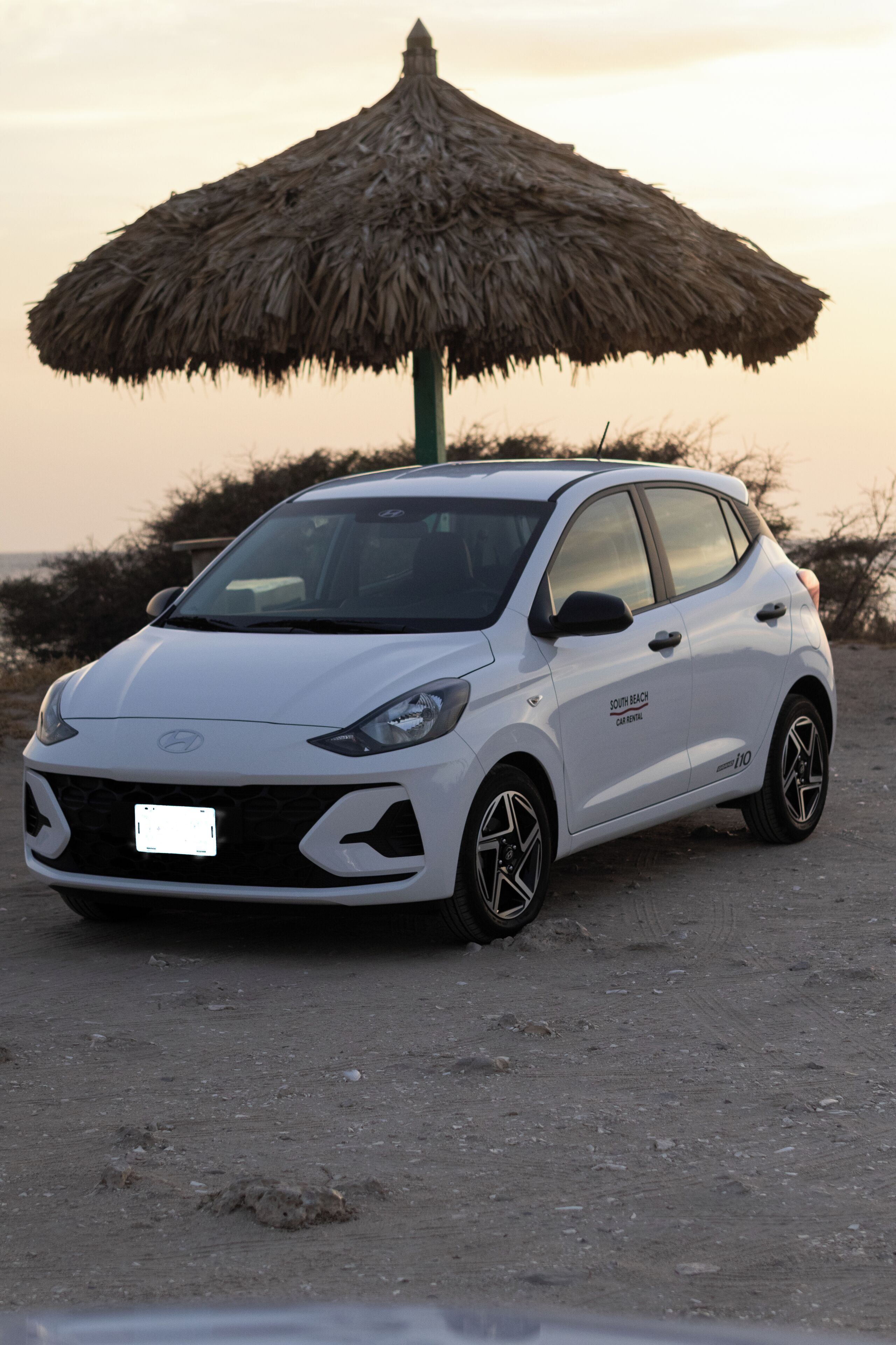A white Hyundai car parked under a thatched umbrella on a dirt path.