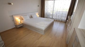 A bedroom with a large bed, wooden flooring, a nightstand with a lamp, and a window with curtains.