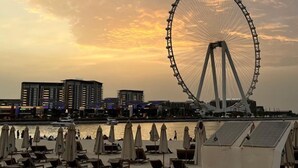 Una spiaggia con lettini e ombrelloni, una ruota panoramica sullo sfondo e edifici lungo la battigia.