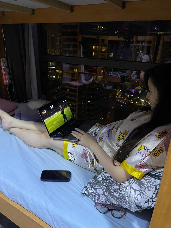A person lying on a bed with a laptop, a smartphone, and a pair of glasses beside them.