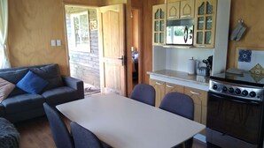 Un salon chaleureux avec un canapé et une table à manger, une cuisine dotée de meubles en bois et d’une cuisinière, et une vue sur l’extérieur grâce à une porte ouverte.