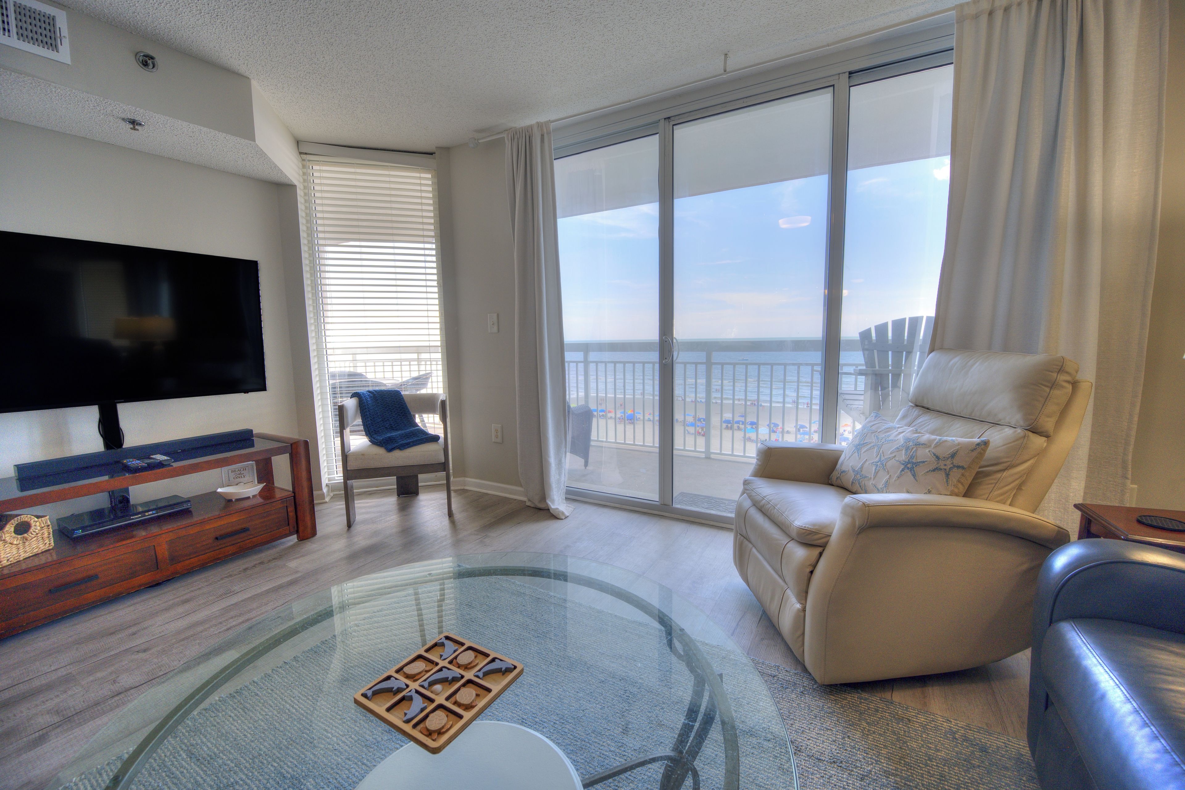 A living room with a flat-screen TV, a glass coffee table, a leather sofa, and a view of the beach through large windows.