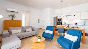 Un salon moderne avec un canapé gris, des fauteuils bleus et une table basse en bois. On aperçoit, au fond, une cuisine avec une table à manger et des chaises.