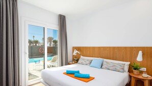 Une chambre d’hôtel avec un grand lit, une tête de lit en bois, une table de chevet avec une lampe et une vue sur une piscine, visible à travers une porte coulissante.
